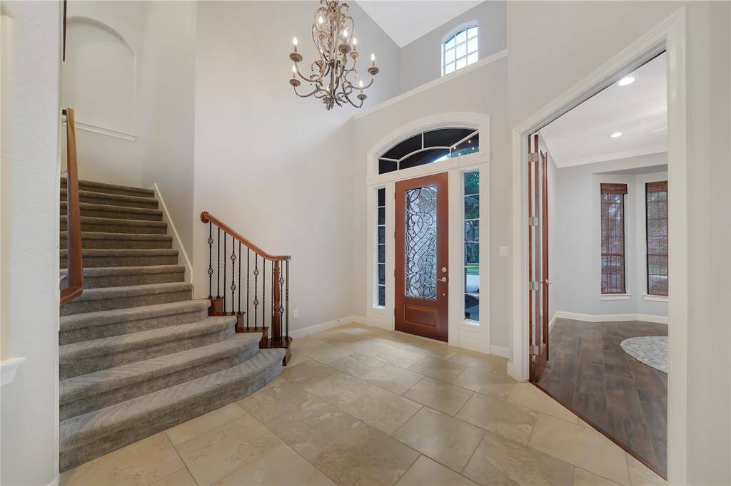 This elegant entryway features a grand staircase with carpeted steps and wrought iron railing, a high ceiling with a chandelier, and a beautiful wooden front door with decorative glass panels. The space is bright with natural light from the large windows, and there's an adjacent room with hardwood floors.