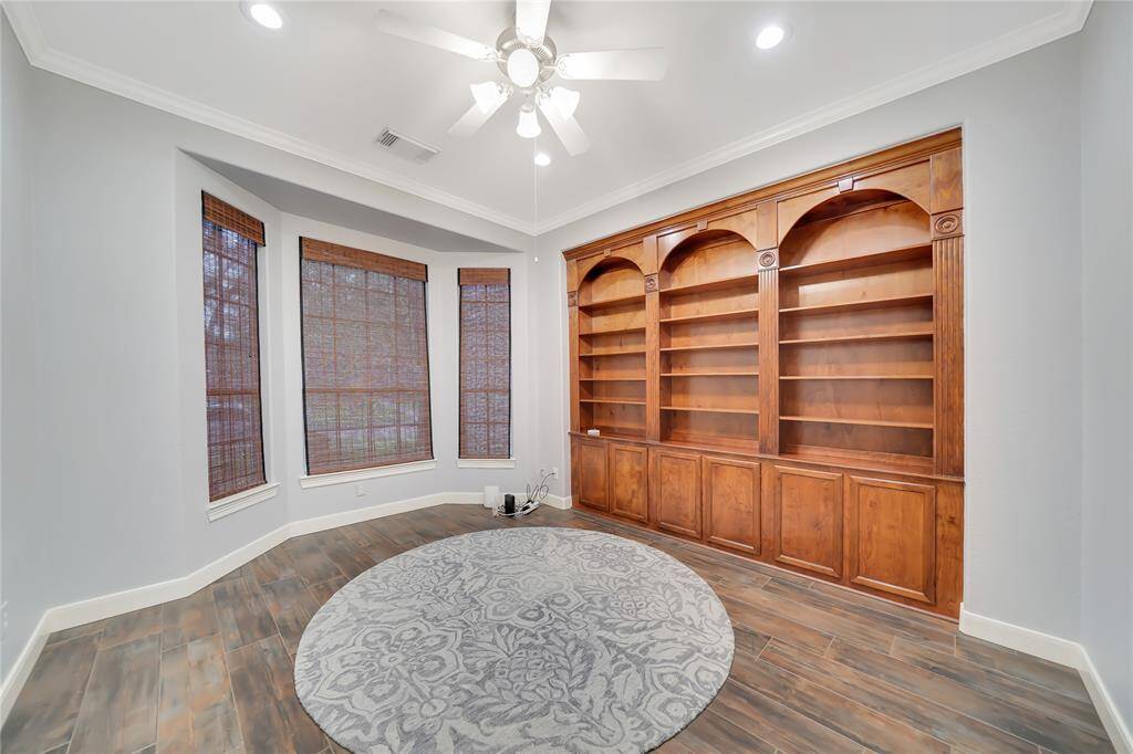 This room features a spacious built-in wooden bookshelf, a large bay window with bamboo shades, and a ceiling fan with recessed lighting. The wood flooring and neutral wall color create a warm, inviting atmosphere. Perfect for a study or reading room.