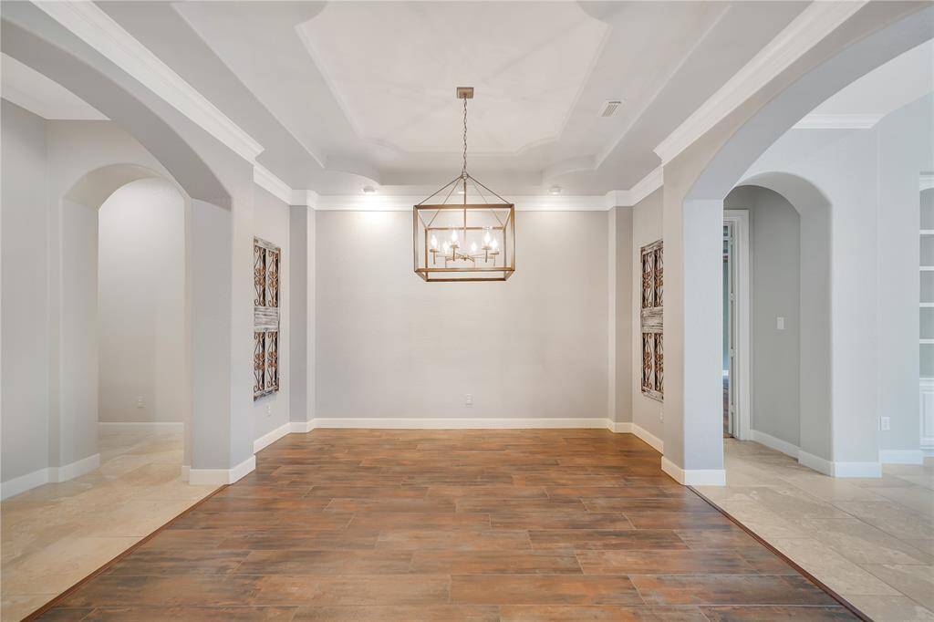 The formal dining features a spacious, elegant design with a mix of wood and tile flooring. It has high ceilings, a modern chandelier, and decorative wall accents, creating a sophisticated ambiance. Arched doorways lead to adjoining spaces, enhancing the open feel.