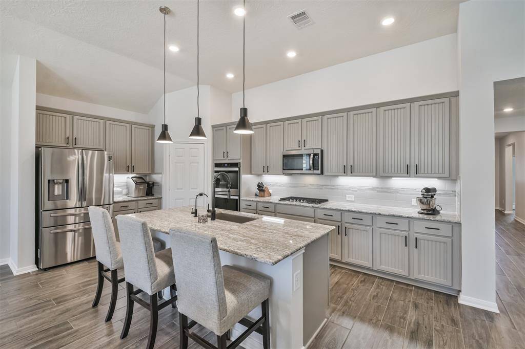 This modern kitchen features sleek gray cabinetry, stainless steel appliances, and a large granite island with seating. The space is well-lit with recessed lighting and pendant lamps, offering a blend of style and functionality.
