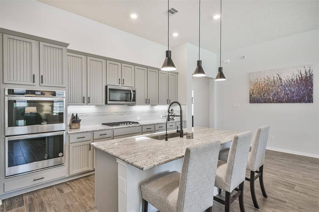 This modern kitchen features sleek gray cabinets, stainless steel appliances, and a spacious island with bar seating. Pendant lighting and an elegant backsplash add a touch of sophistication, while the open layout enhances functionality.