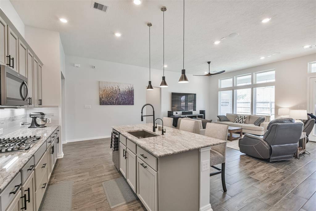This modern open-concept living space features a spacious kitchen with granite countertops, pendant lighting, and a large island. It flows into a bright living area with large windows, tile floors.