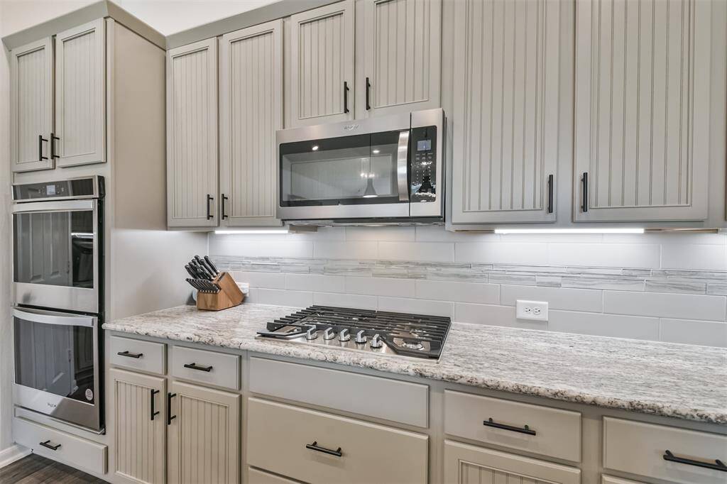 This modern kitchen features sleek cabinetry with vertical paneling, stainless steel appliances including a double oven and microwave, a gas cooktop, and a stylish granite countertop. The backsplash combines subway and accent tiles for a contemporary look.