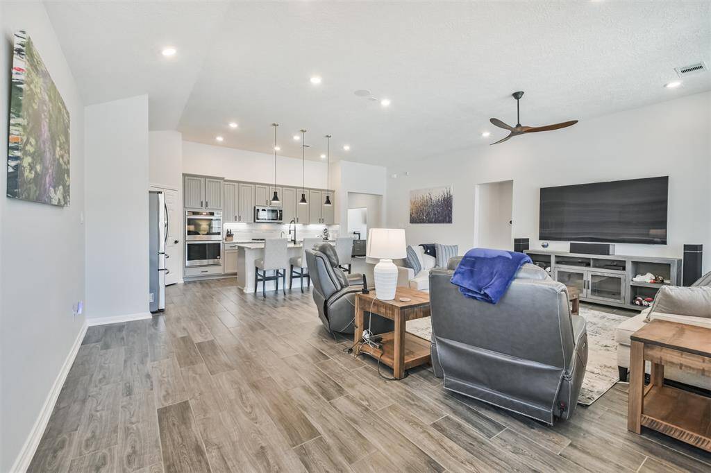 This spacious, modern living area features an open-concept design with a sleek kitchen, complete with stainless steel appliances and a large island. The room is bright and airy, with wood-look tile flooring offering a comfortable and inviting atmosphere.