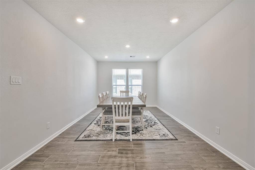 This photo showcases a bright, elongated dining room with neutral walls, recessed lighting, and wood-look flooring. It features a large dining table with chairs and a decorative rug, leading to a window at the far end.