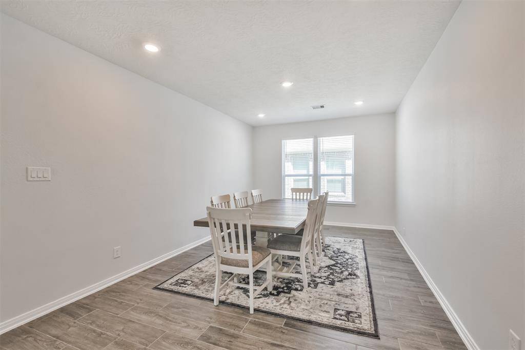 This photo showcases a spacious, well-lit dining room with neutral walls and wood-style tile flooring. It features a large table with chairs, set on a patterned area rug, and natural light streaming through double windows.