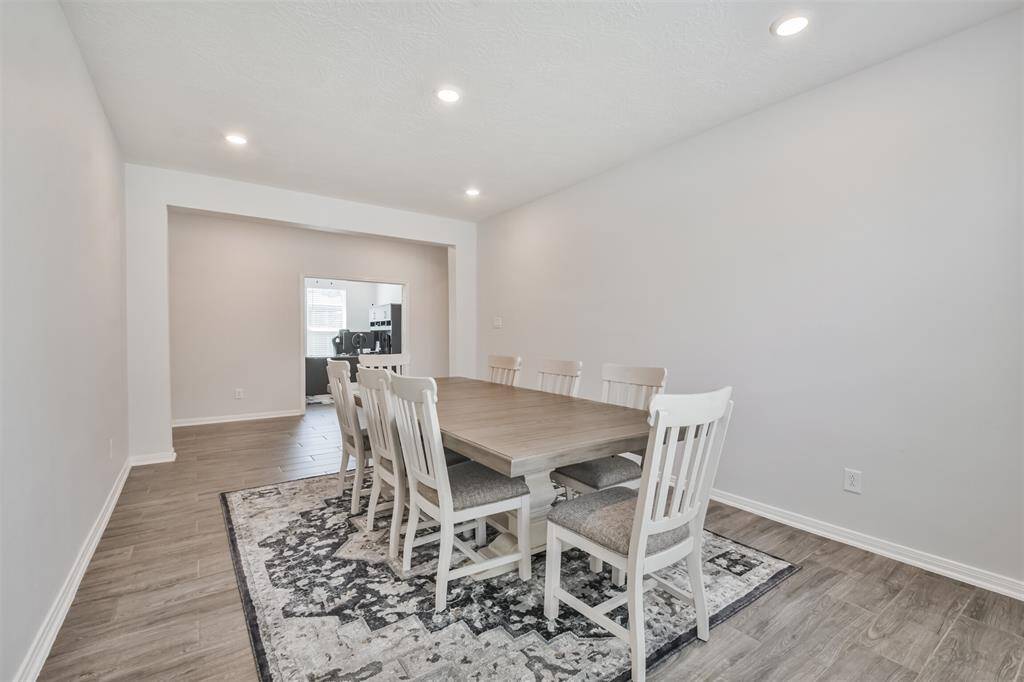 This photo shows a spacious dining room the room has recessed lighting and connects to another area, likely a kitchen or living space, with natural light coming through a window.