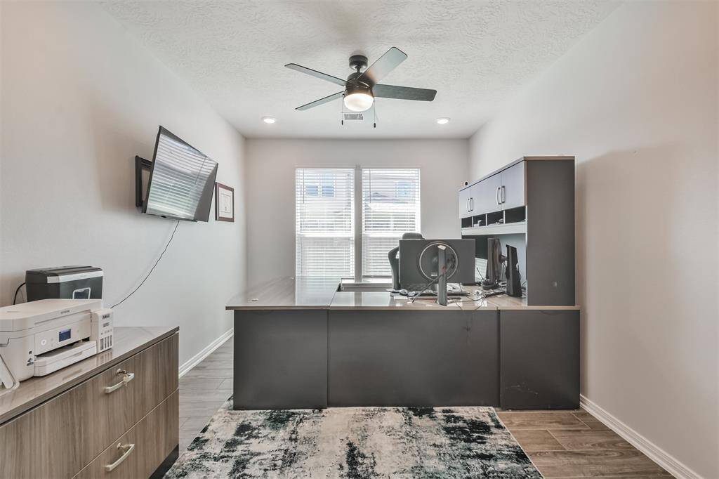 This photo showcases a modern home office with a large desk, ergonomic chair, and ample storage. It features a ceiling fan, wall-mounted TV, and a window allowing natural light. The room has a sleek, professional look with neutral tones and wood flooring.