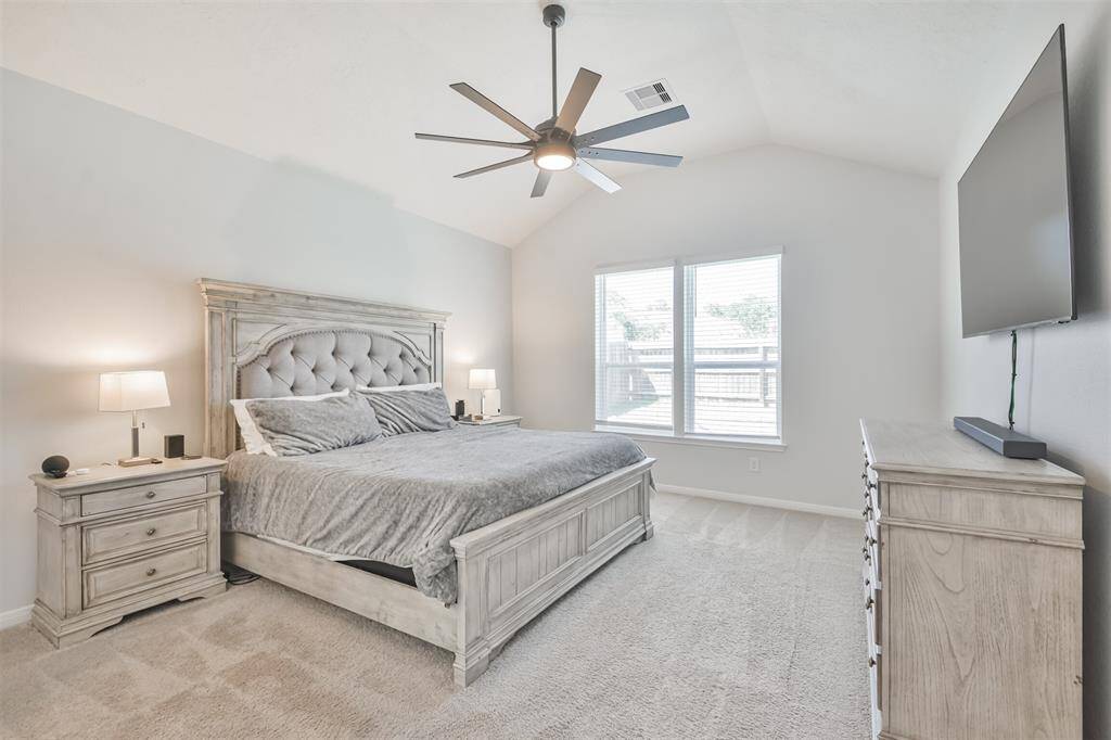 This bedroom is spacious and bright, featuring a vaulted ceiling with a modern ceiling fan. It includes a large bed with a tufted headboard, matching nightstands, and a dresser. Natural light streams through two sizable windows, enhancing the room's airy feel. A mounted flat-screen TV adds a contemporary touch.