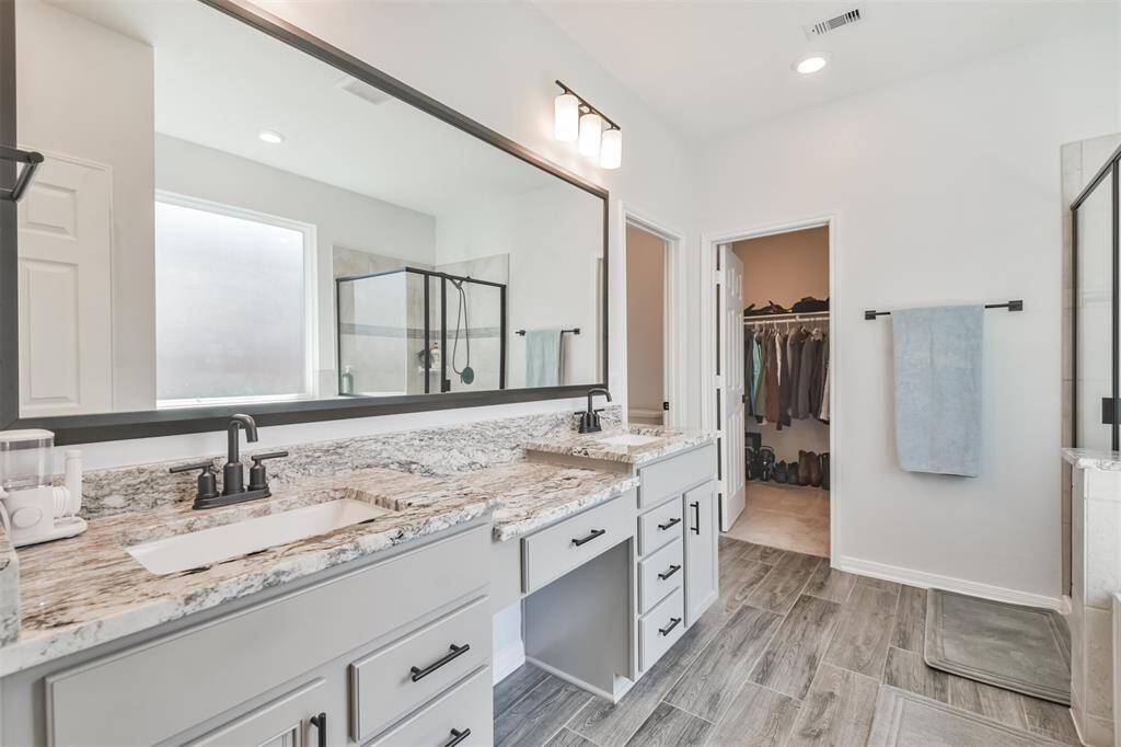 This bathroom features a spacious double vanity with granite countertops and modern fixtures, a large mirror, and a walk-in shower with glass doors. The neutral color palette and wood-look tile flooring create a sleek, contemporary feel. A walk-in closet is accessible directly from the bathroom.