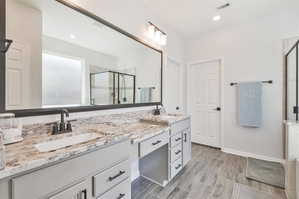 This spacious bathroom features a double vanity with granite countertops, modern fixtures, and ample storage. It includes a large mirror, a glass-enclosed shower, and stylish tile flooring, offering a clean and contemporary feel.
