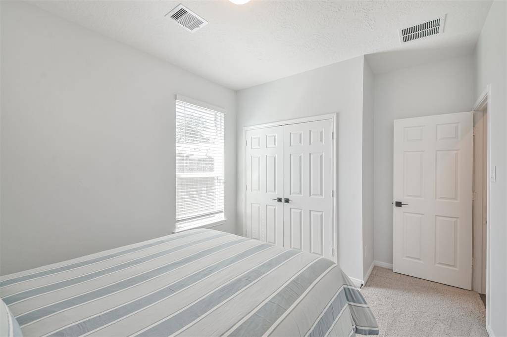 This photo showcases a bright, cozy bedroom with neutral walls and carpet. It features a window with blinds for natural light, and ample closet space with double doors.