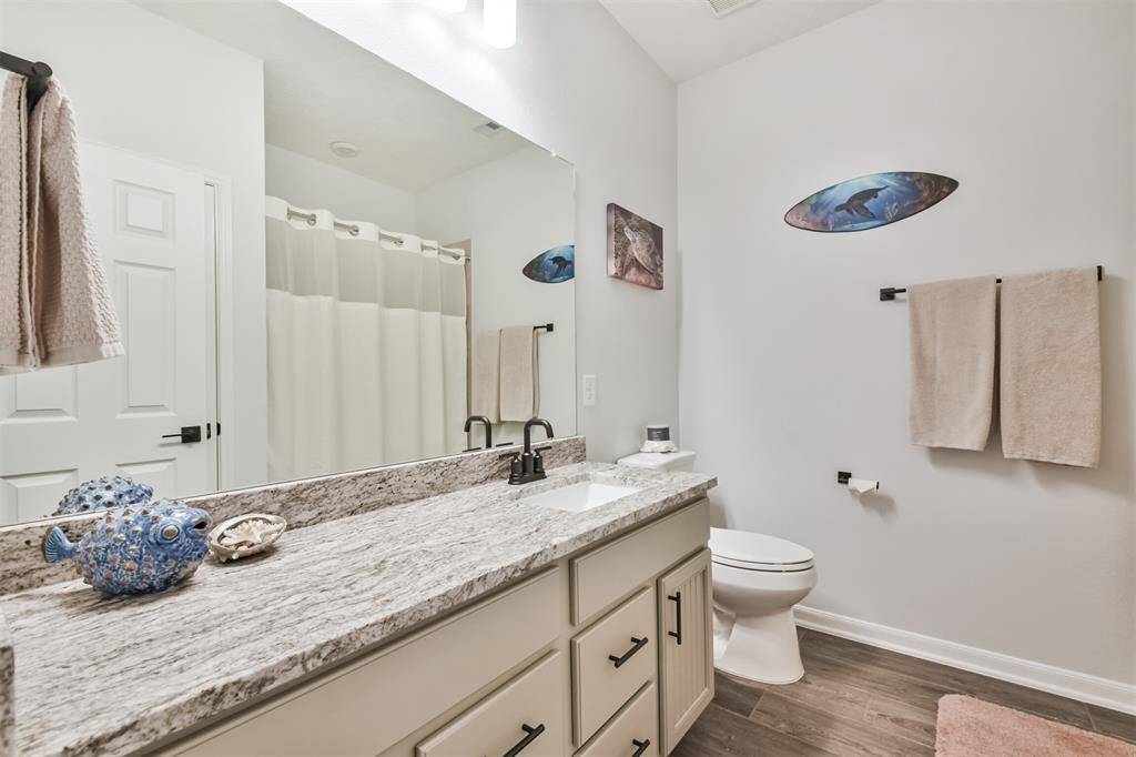 This bathroom features a modern design with a granite countertop, undermount sink, and sleek fixtures. It includes a shower with a curtain, neutral wall colors, and decorative accents for a cozy feel. The floor is finished with wood-like tiles, enhancing the overall aesthetic.
