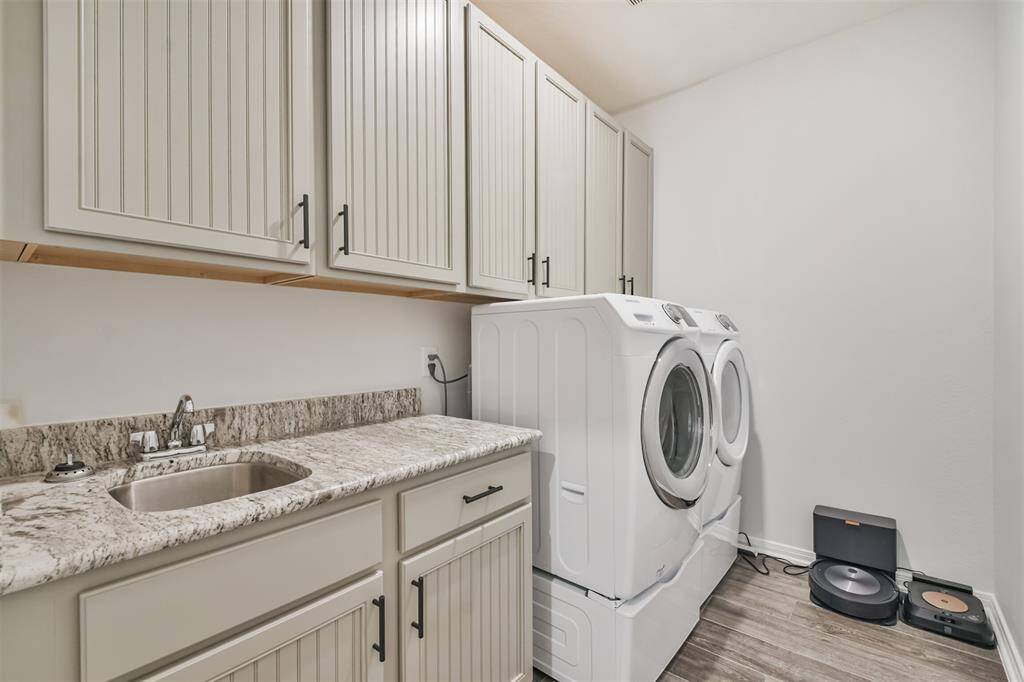 This photo shows a modern laundry room with white cabinetry, granite countertops, a stainless steel sink, and a washer and dryer. The space also features wood-look flooring and ample storage.