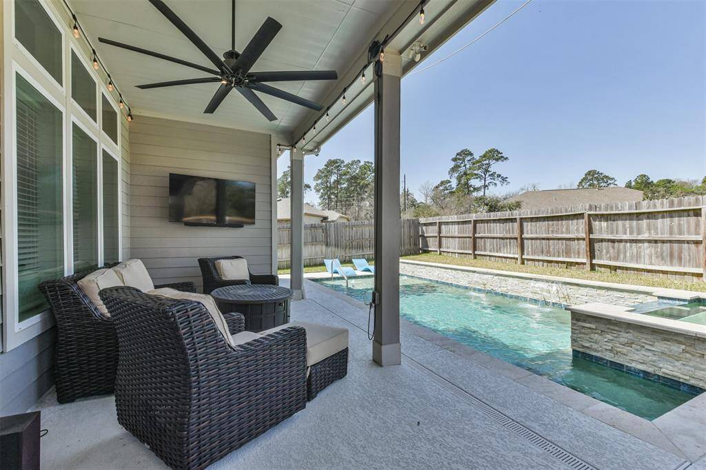 This photo showcases a modern outdoor patio with lighting, a ceiling fan, and a wall-mounted for a TV. It overlooks a sleek pool with a stone waterfall feature, all enclosed by a wooden privacy fence, offering a relaxing and private entertainment space.
