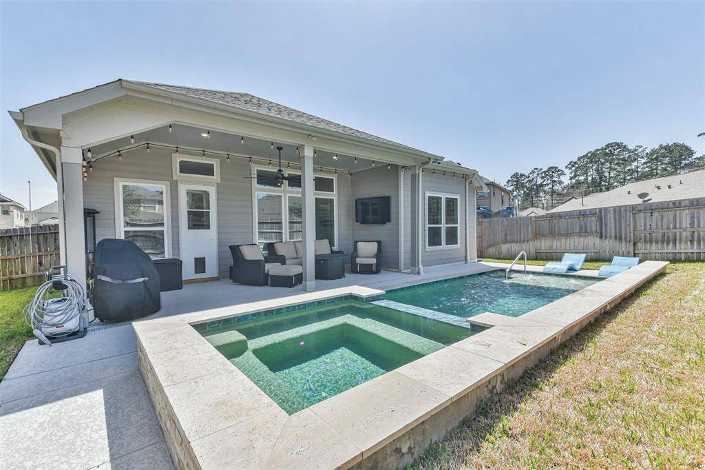 Backyard with a pool and hot tub, a covered patio with seating, and an outdoor TV. The area is enclosed by a wooden fence, providing privacy and a cozy outdoor living space.