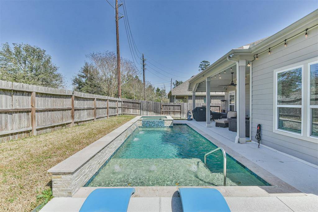 This photo features a backyard with a modern in-ground pool, complete with a small waterfall feature. The area is enclosed by a wooden fence, providing privacy. There's a covered patio attached to the house, offering outdoor seating and relaxation space. The setting is ideal for enjoying sunny days and outdoor entertaining.