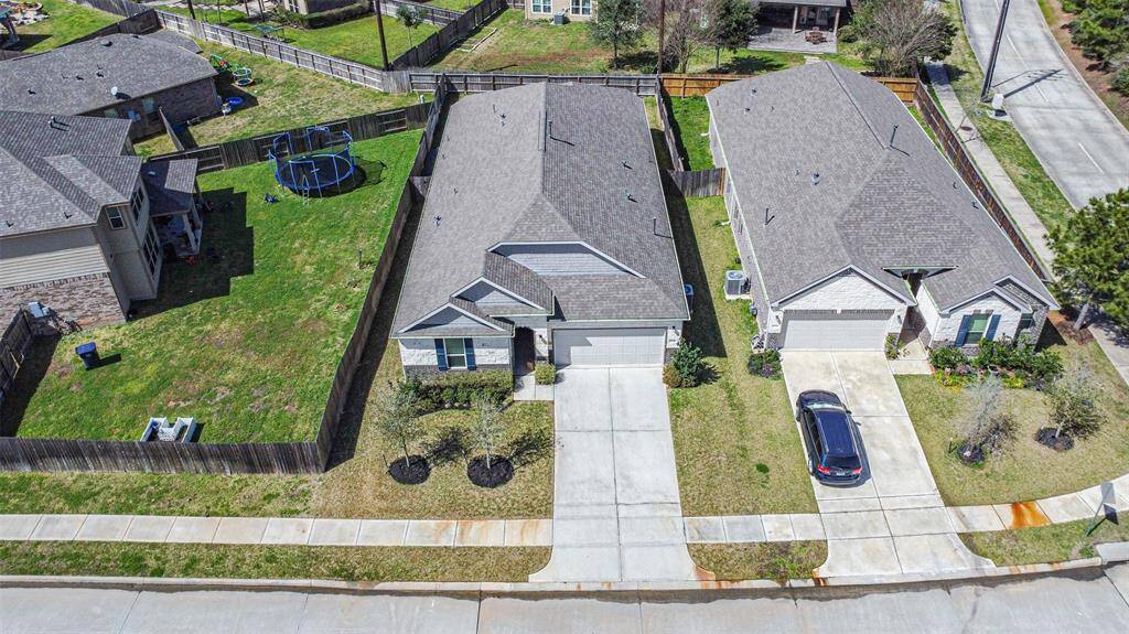 This aerial view shows a suburban neighborhood with two single-story homes featuring gray roofs and well-maintained front lawns. Each house has a driveway leading to a two-car garage. The backyards are fenced, providing privacy, and one includes a trampoline. The street is lined with sidewalks and mature trees, creating a welcoming environment.