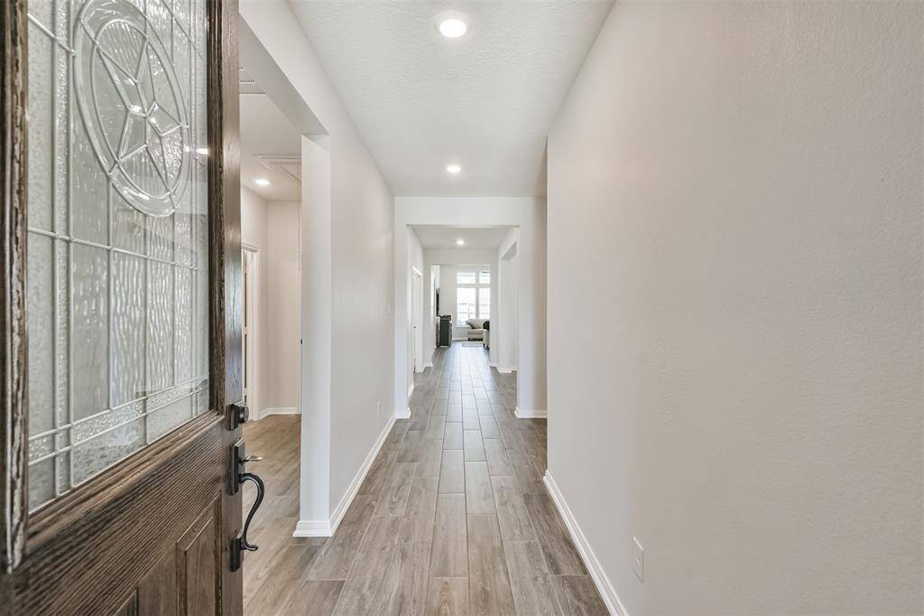 This photo shows a bright, welcoming entryway with wood-look tile flooring, neutral walls, and recessed lighting, leading toward a living area. The entry door features decorative glass.