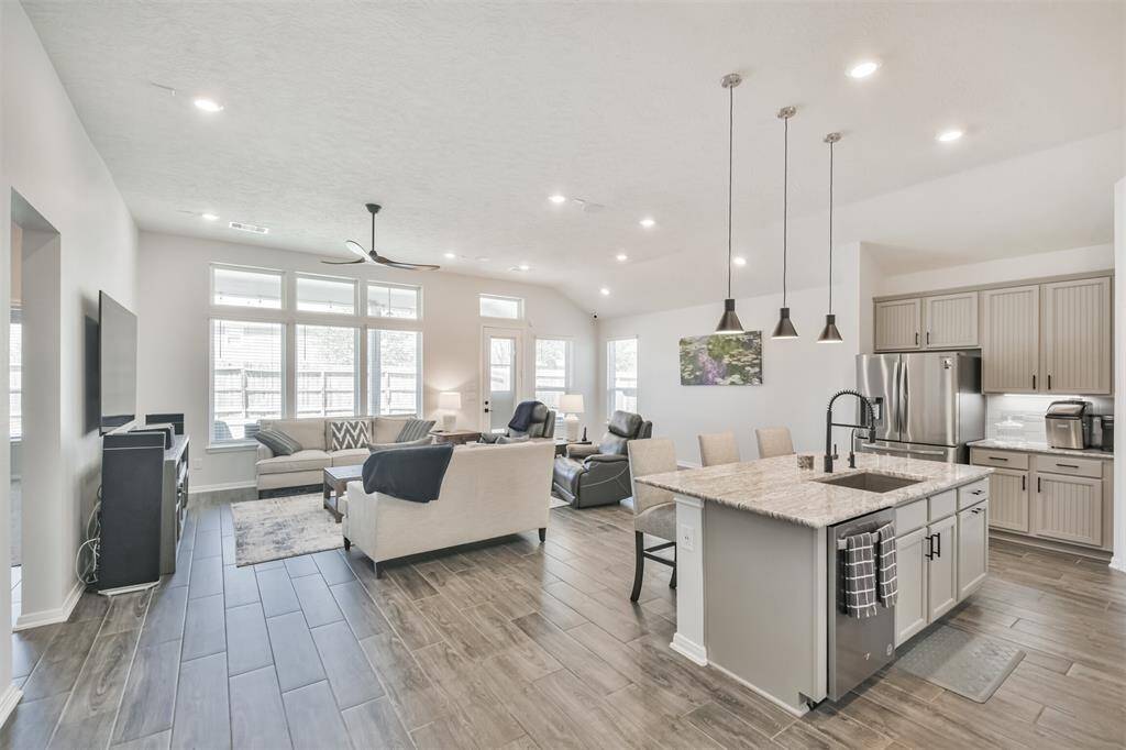 This photo showcases a bright, open-concept living area with large windows, modern lighting, and a ceiling fan. It features a spacious kitchen with an island, granite countertops, stainless steel appliances, and ample cabinetry. The adjoining living room includes a cozy seating area, creating a welcoming and functional space.