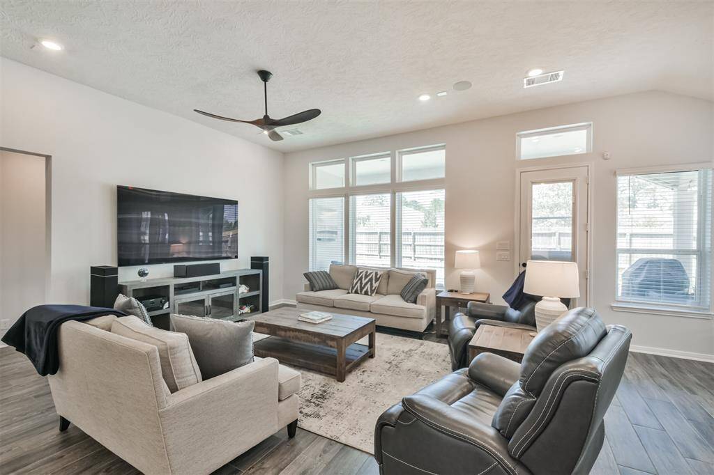 This living room is spacious and bright, featuring large windows, modern furnishings, and a ceiling fan. The neutral palette and ample seating create a welcoming atmosphere, perfect for relaxation and entertainment.