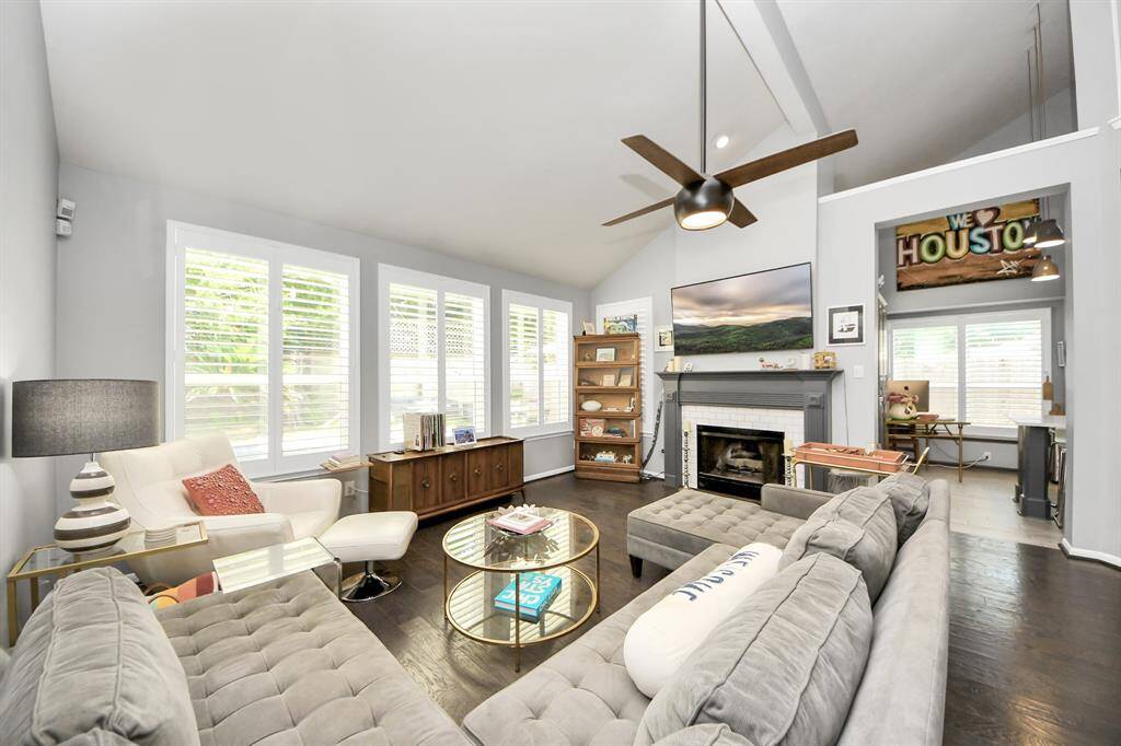 This inviting living room features vaulted ceilings, a sleek modern fan, and a cozy fireplace with a custom mantel. Large windows fill the space with light, creating a bright and airy retreat for family gatherings.