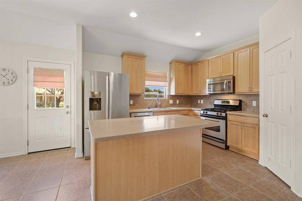 This kitchen features modern stainless steel appliances, light wood cabinetry, and a central island. It has ample natural light, a tile floor, and a convenient door to the backyard.