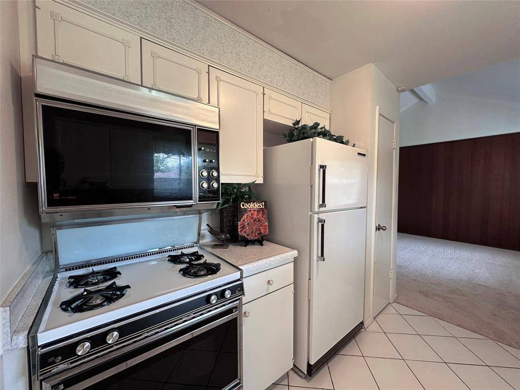 This kitchen features vintage appliances, including a gas stove, oven and fridge, with white cabinetry and countertops. The flooring is tiled, and the space has a classic, retro feel - all open to the inviting and freshly carpeted living room.