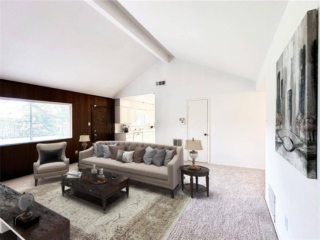Bright, spacious living area with vaulted ceiling, large window for natural light, and an adjacent kitchen. Carpet throughout installed in May 2025 and a mix of wood create a cozy atmosphere.  Image Virtually Staged.