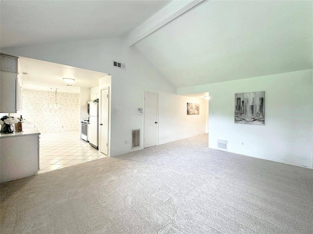 Bright and spacious living area with vaulted ceilings and fresh carpet throughout. The open layout leads into a tiled kitchen and breakfast area, featuring white cabinetry, creating a clean and inviting family atmosphere.