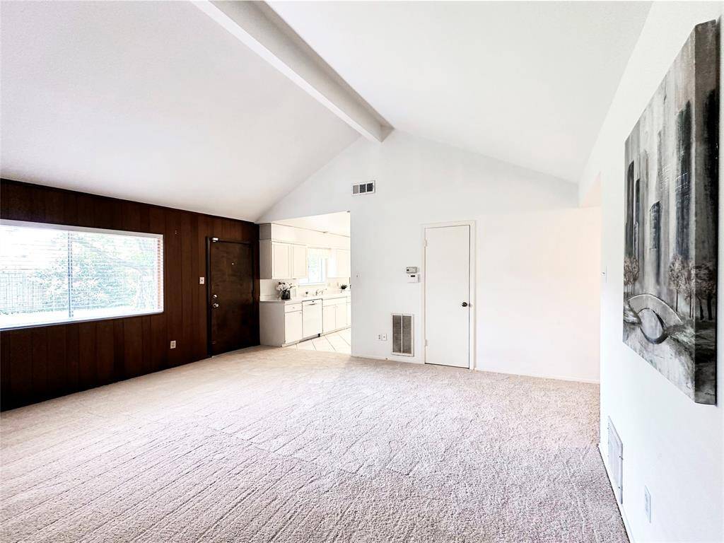 Bright, spacious living area with vaulted ceiling, large window for natural light, and an adjacent kitchen. With fresh carpet throughout and a mix of wood create a cozy atmosphere.
