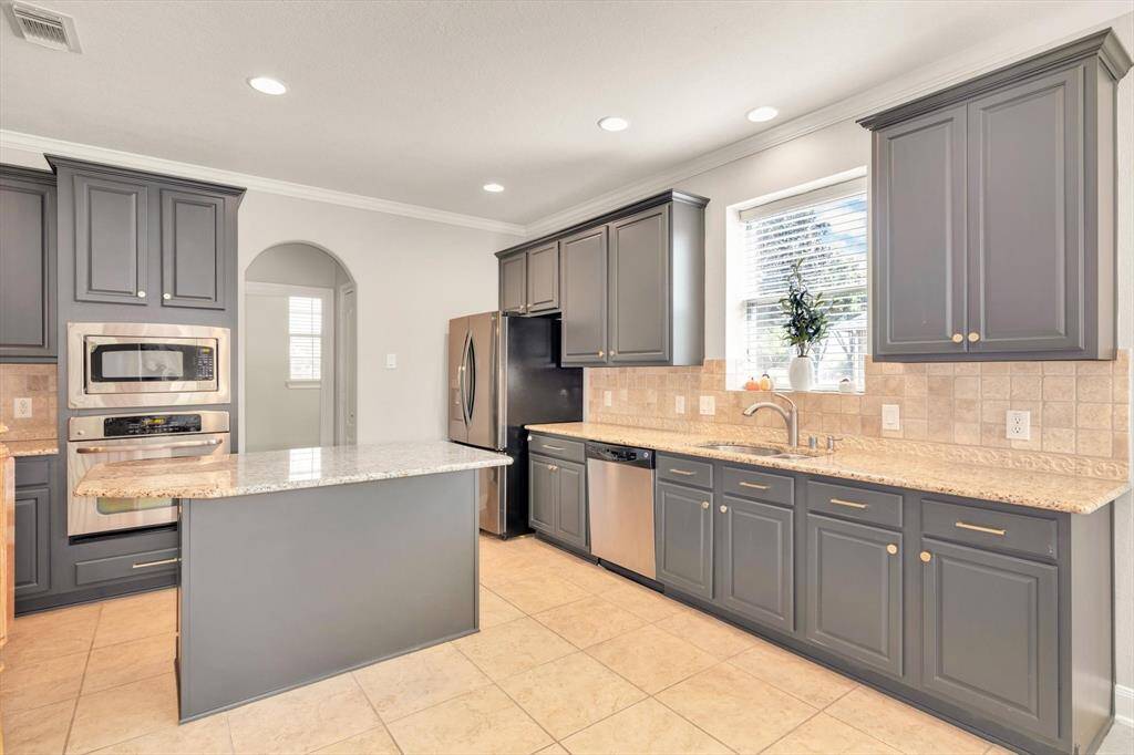 This modern kitchen features sleek gray cabinets, granite countertops, stainless steel appliances, and a central island. It has ample natural light and a tiled backsplash, offering a clean and inviting space.