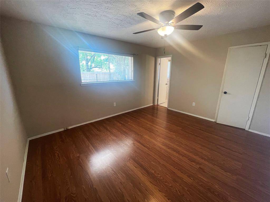 This photo shows a spacious bedroom with warm wood flooring, a ceiling fan, and neutral-colored walls. It features a large window letting in natural light and has a white door leading to an adjacent room or bathroom.