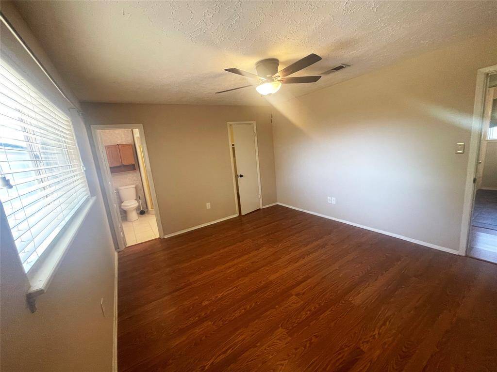 This is a bright bedroom featuring wood flooring, a ceiling fan, and a window with blinds. It has a door leading to an en-suite bathroom.
