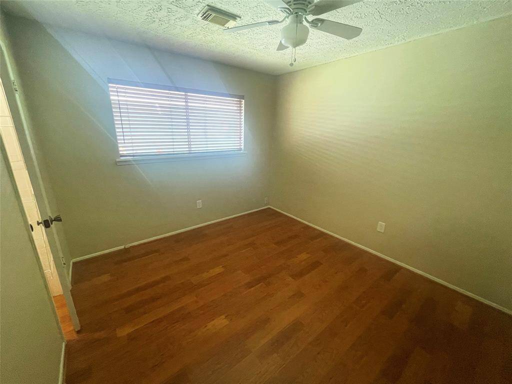 This photo shows a small, empty room with wooden floors and a ceiling fan. Natural light enters through a window with blinds, and the walls are painted a neutral color.