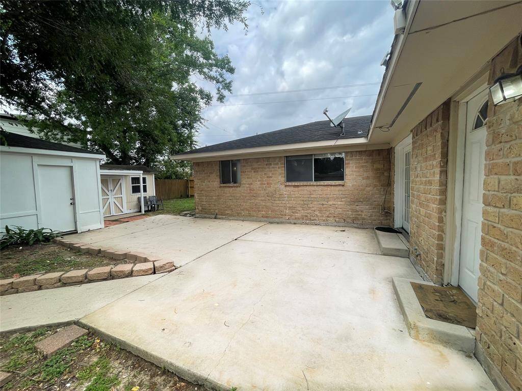 This photo showcases a spacious backyard area with a concrete patio, perfect for outdoor activities. The setting includes a brick exterior, a storage shed, and mature trees providing shade.