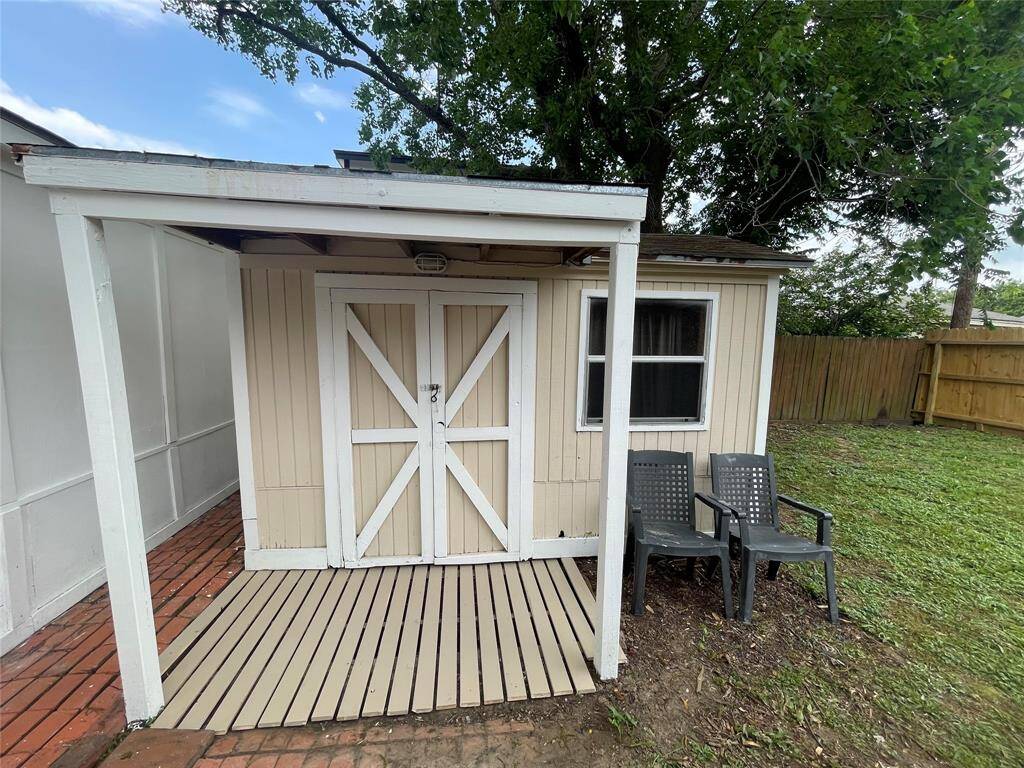 This photo shows a small backyard shed with beige siding and a white door, featuring a covered entryway. There are two black chairs on a grassy area, and a wooden fence runs along the property line. A large tree provides shade in the background.