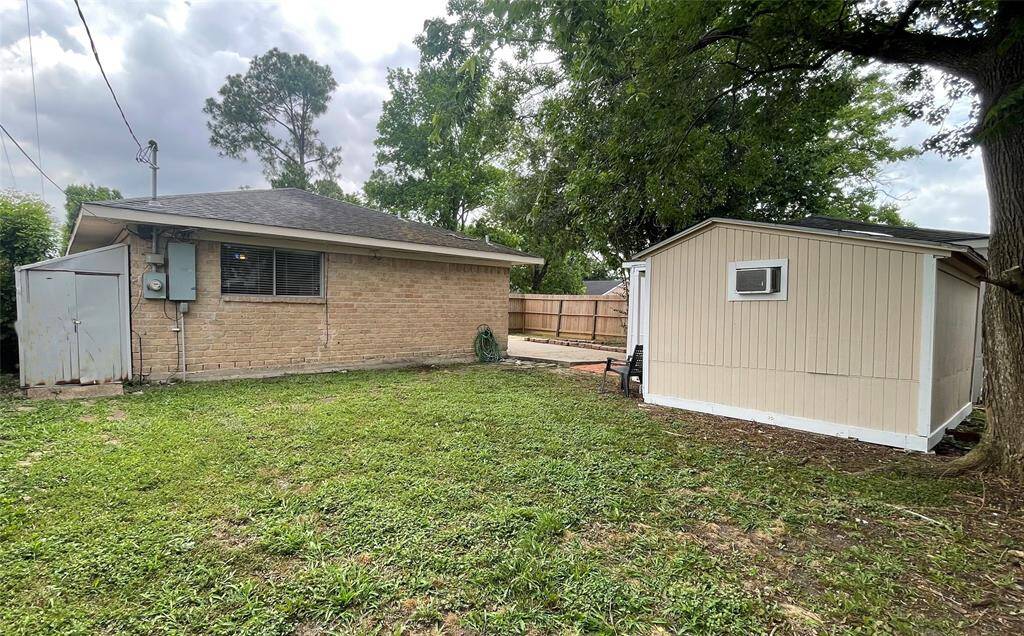This photo shows a backyard with a brick house and a separate shed. The yard is grassy with a few trees, and there's a wooden fence providing privacy. A small paved area is adjacent to the house.