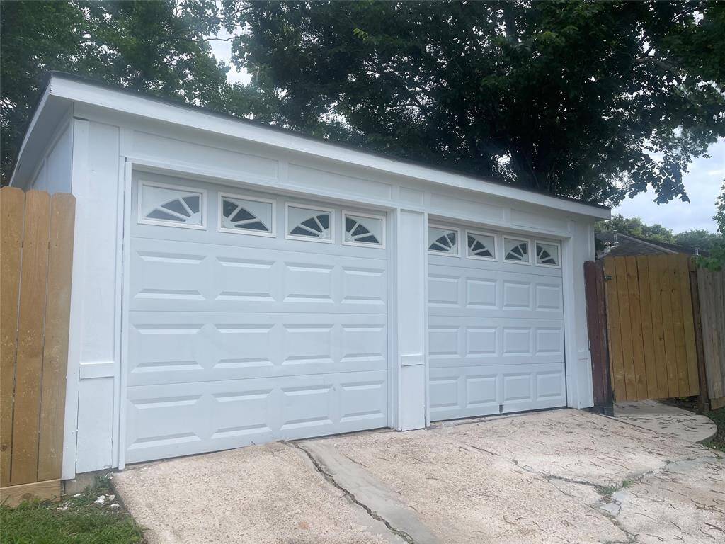 This photo features a clean, white two-car garage with decorative windows. The garage doors are set in a sturdy structure, surrounded by a wooden fence and shaded by trees, providing a secure and private parking area.