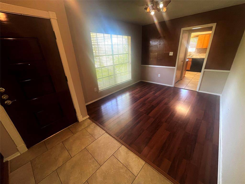 This photo showcases a bright entryway with a mix of tiled and hardwood flooring. It features a large window with natural light, modern lighting fixtures, and an open doorway leading to a kitchen area.
