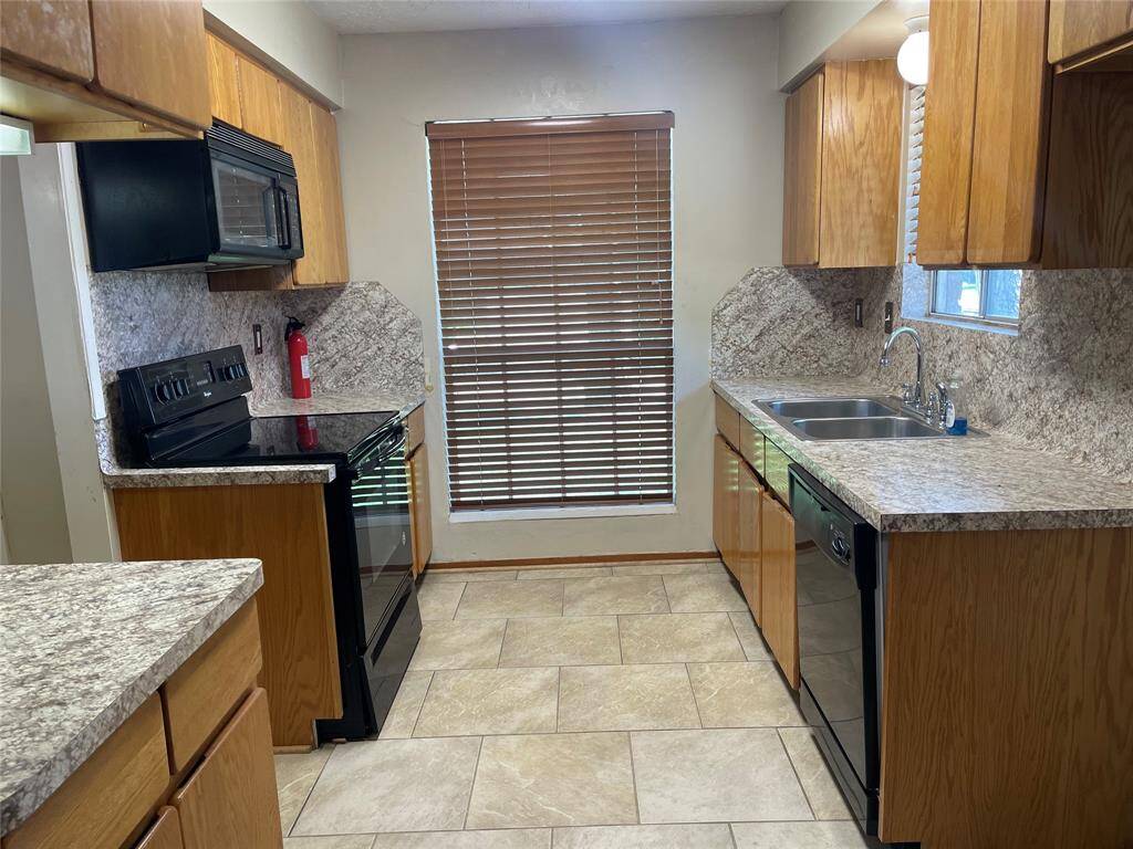 This kitchen features wood cabinetry, granite countertops, and modern black appliances. There's a window with blinds for natural light, tiled flooring, and a double sink, providing a practical and inviting space.