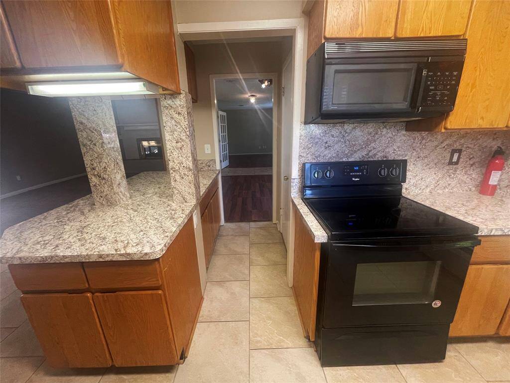 This kitchen features granite countertops, wooden cabinetry, a black electric stove, and a built-in microwave. The open layout provides a view into an adjacent room, enhancing connectivity and space.