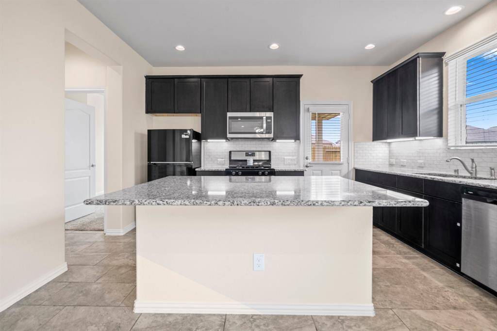 Kitchen features sleek black cabinetry, granite countertops, and a white subway tile backsplash. It includes a built-in dishwasher and a stainless steel sink with a modern faucet. Natural light flows in through a nearby window, complementing the bright and clean aesthetic.