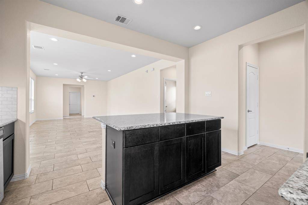Spacious, open-concept kitchen and living area with light tile flooring, neutral walls, and a granite-topped island. Recessed lighting and large windows provide ample natural light, enhancing the airy feel of the space.