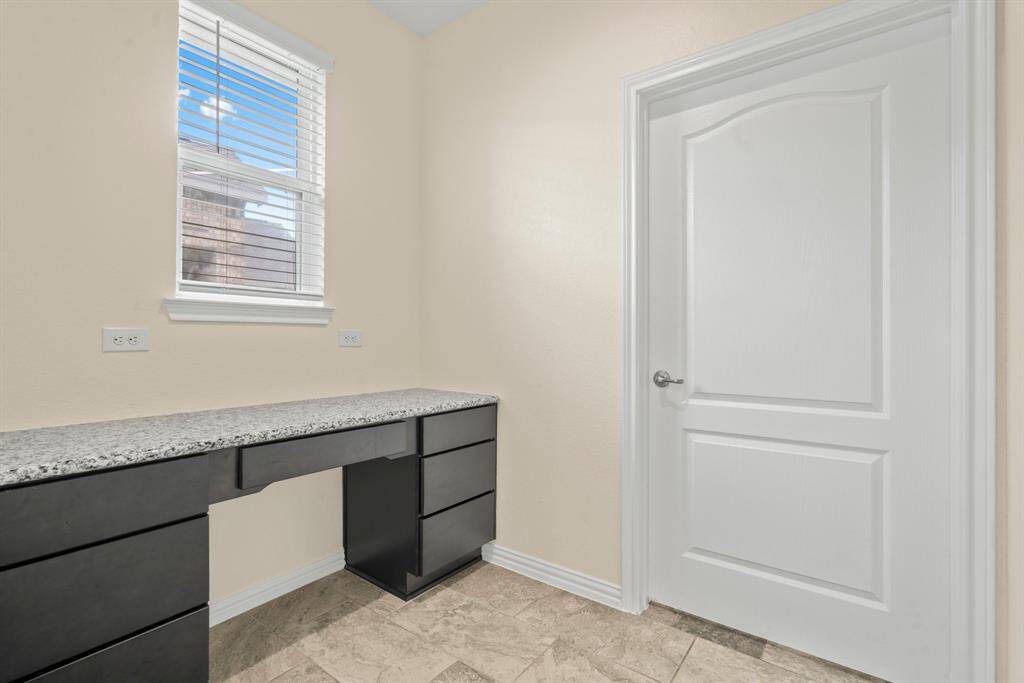 Functional workspace with a built-in black granite desk and drawers. The room has a window for natural light, beige walls, tile flooring, and a white door. Perfect for a home office or study nook.
