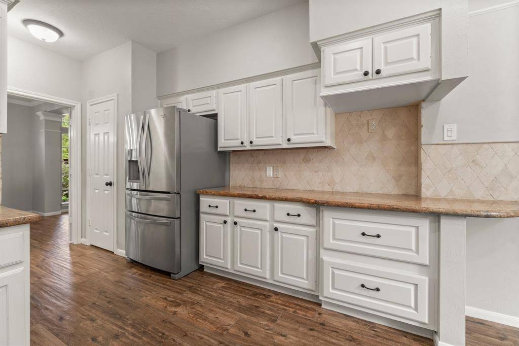 This kitchen cabinetry features dark handles, granite countertops, and a stainless steel refrigerator. The backsplash has a neutral tile design, and the space opens to a room flooded ample natural light and enough space for multiple cooks.
