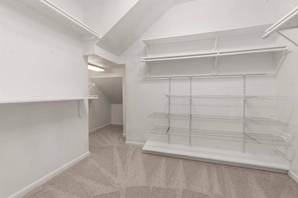 This spacious primary walk-in closet features ample shelving and hanging space, with carpeted flooring and bright lighting, perfect for organized storage.