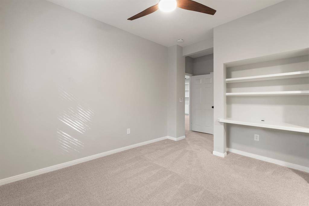 This room features soft neutral walls and carpet, a ceiling fan, built-in shelves, and a desk area. Perfect for an office. study space, or bedroom.