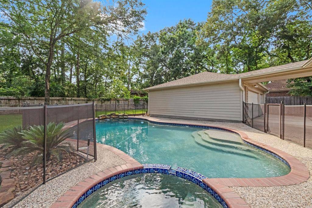 This photo showcases a spacious backyard with a curvy swimming pool and attached spa, surrounded by a safety fence. The area is bordered by trees and a wooden fence, providing privacy. The garage is visible in the background, adding a practical element to the outdoor space.
