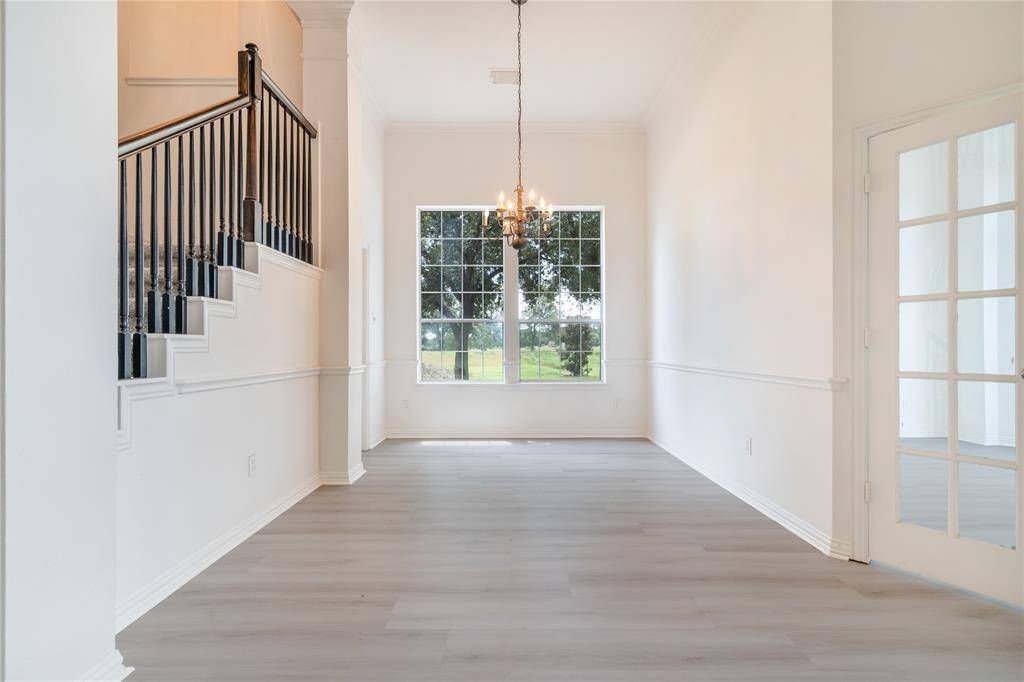 This home showcases a spacious, bright hallway with high ceilings, elegant hardwood flooring, and a grand staircase with dark railings. Large windows provide natural light and a view of the greenery outside. This home also features classic wainscoting and a chandelier, adding a touch of sophistication.